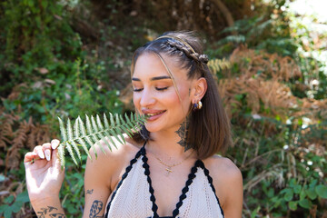 Portrait of a beautiful young woman with tattoos on her arms and neck holding a green fern leaf in her hand and biting it. The woman is smiling and enjoying herself.