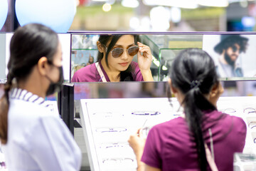 A young woman tries sunglasses at the mirror in an optical shop with the assistance of a saleswoman