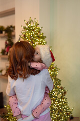A girl in a New Year's hat hugs her mother on New Year's at home; a woman celebrates Christmas with her daughter, congratulates the child and gives her a gift.
