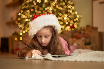 Cute little child girl writing letter to Santa Claus or writing dreams of a gift with near Christmas tree. Merry Christmas and Happy New Year!