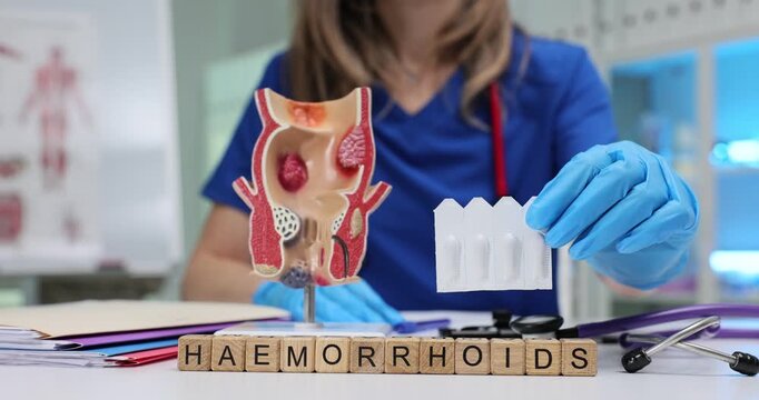 Wooden blocks spell word Haemorrhoids near anorectal model display. Proctology doctor holds suppository blister explaining haemorrhoids treatment