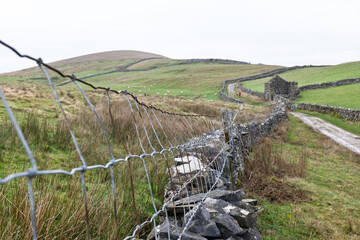 Agricultural wire mesh fencing attached to the top of a dry stone wall leads off into a Yorkshire Dales landscape on a wet overcast day.