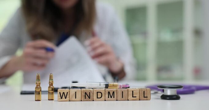 Wooden blocks spell word Windmill on clinical desk with tools and ampoules. Research woman fills documents recording windmill study note carefully