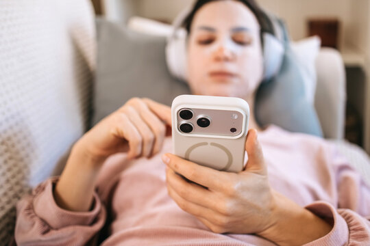 Young woman with headphones relaxing on a couch, using smartphone to browse content, showcasing modern lifestyle and technology in a cozy home environment