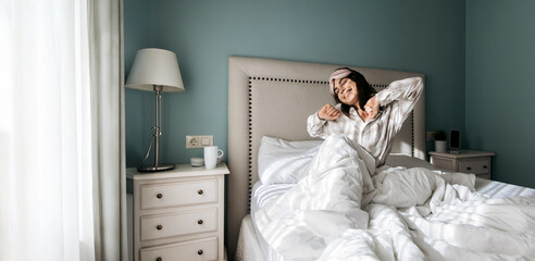 Young woman in cozy pajamas stretches joyfully in a bright bedroom with soft bedding and a stylish lamp, capturing a serene morning routine and relaxation atmosphere
