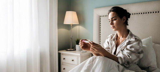 Woman in cozy bedroom, sitting on bed, holding pregnancy test, with soft natural light illuminating the scene, conveying anticipation and emotional moment of discovery