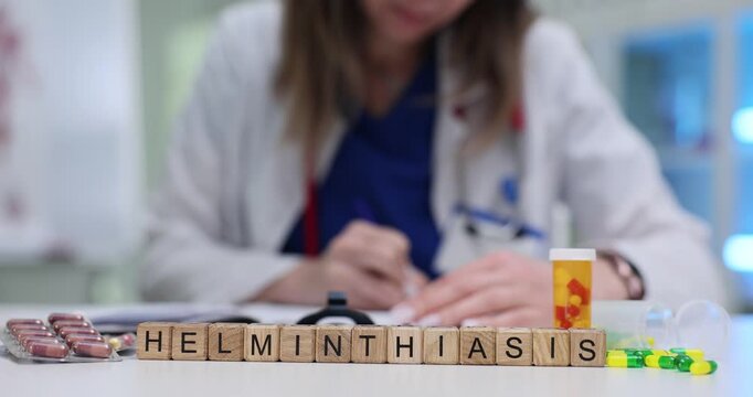 Wooden blocks spell word Helminthiasis near stethoscope on desk. Doctor woman writes prescription choosing treatment for helminthiasis at appointment