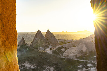 View of sunbeams bursting over the horizon, illuminating the rugged, rocky landscape in warm hues, captured through a natural stone frame, Goreme, Cappadocia, Turkey.