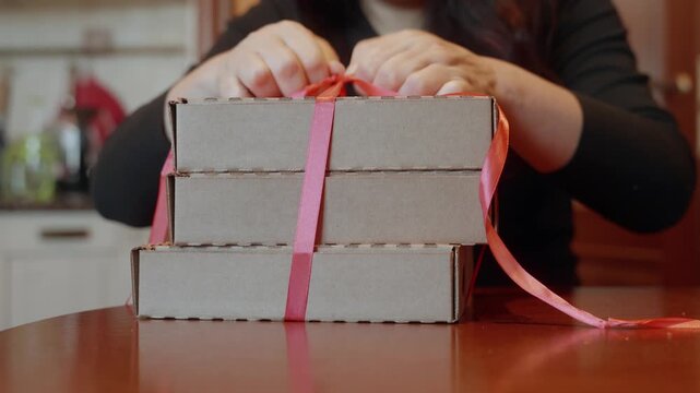 Close up of woman in black shirt with red nails untying pink ribbon bow on stack of cardboard pizza boxes at wooden table, unpacking food delivery.
