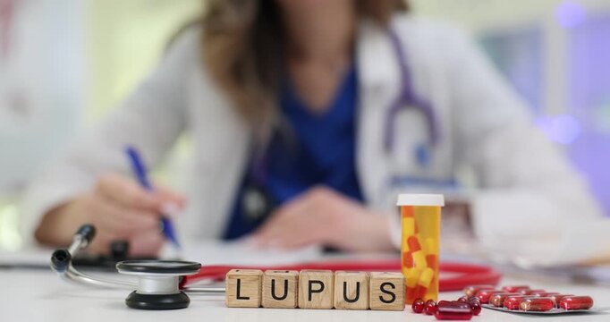 Wooden blocks spell word Lupus near stethoscope tray and pill bottle. Rheumatology doctor writes notes monitoring lupus symptoms with treatment