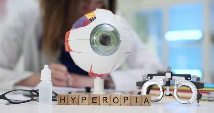 Wooden blocks spell word Hyperopia near giant eyeball model on desk. Ophthalmology woman writes chart testing hyperopia with trial frame at table