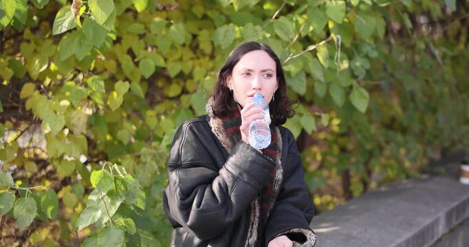 Young woman sits on parapet holding plastic water bottle in park. Female drinks cool water from bottle taking short relaxing rest at sunny weather