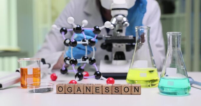 Wooden blocks spell word Oganesson near molecular lattice on table. Laboratory person adjusts model studying oganesson properties during experiment
