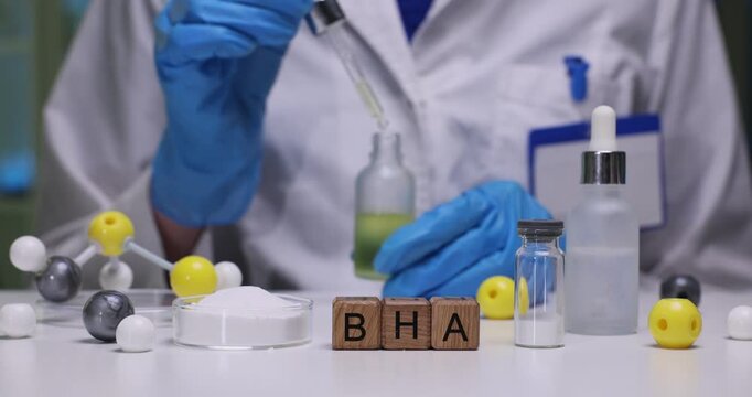 Technician holds glass bottle containing golden oil. Worker tilts bottle slightly to see consistency of oil. BHA abbreviation made from wooden cubes