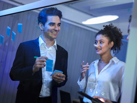 Diverse business professionals executives, collaborating on strategy. Brainstorming, writing on a transparent glass board. Using digital tablets for successful, corporate development and innovation