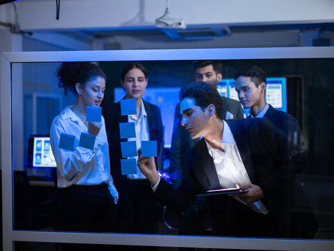 Diverse team of business professionals collaborating on a glowing digital smart screen in the office. Writing strategy with sticky note on glass board. Demonstrating innovative brainstorming