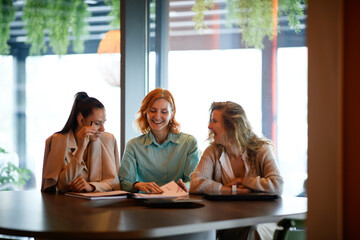 Three businesswomen collaborate at a table in a bright office during the daytime, discussing business proposals and sharing a lighthearted moment