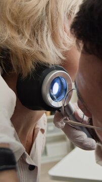 Vertical closeup shot of doctor in white gloves examining neck of blonde female patient with dermatoscope checking for suspicious moles during appointment in dermatology clinic