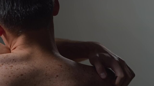 Closeup rear view shot of unrecognizable adult man scratching itchy skin on bare back on minimal dark background in studio