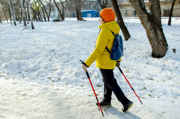 Senior woman in yellow coat doing Nordic walking along snowy path in city park