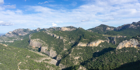 Aerial view of a mountain range with trees and cliffs under a blue sky with white clouds above it all