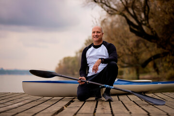 Sporty active senior man sitting on the jetty by the river next to his kayak