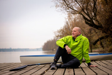 Sporty active senior man sitting on the jetty by the river next to his kayak