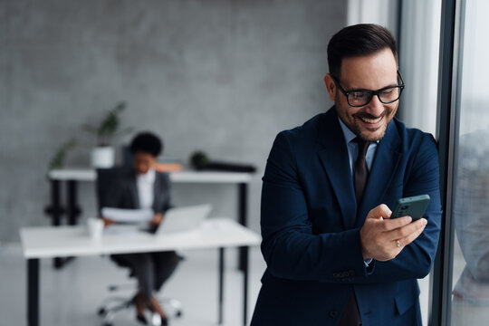 A businessman in a modern office standing near a window and using his smartphone and in the background, an African businesswoman is seated at a desk working on a laptop
 - Powered by Adobe