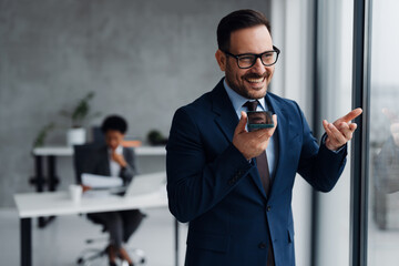 A businessman in a modern office standing near a window and using his smartphone and in the background, an African businesswoman is seated at a desk working on a laptop
