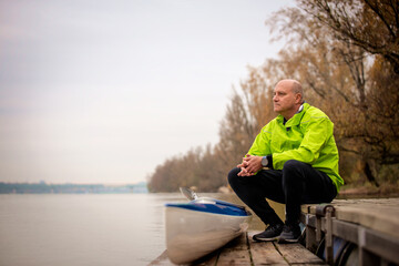 Sporty active senior man sitting on the jetty by the river next to his kayak