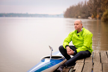 Sporty active senior man sitting on the jetty by the river next to his kayak