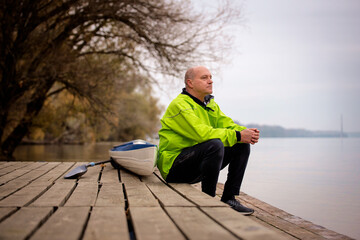 Sporty active senior man sitting on the jetty by the river next to his kayak
