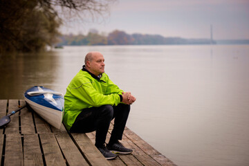 Sporty active senior man sitting on the jetty by the river next to his kayak