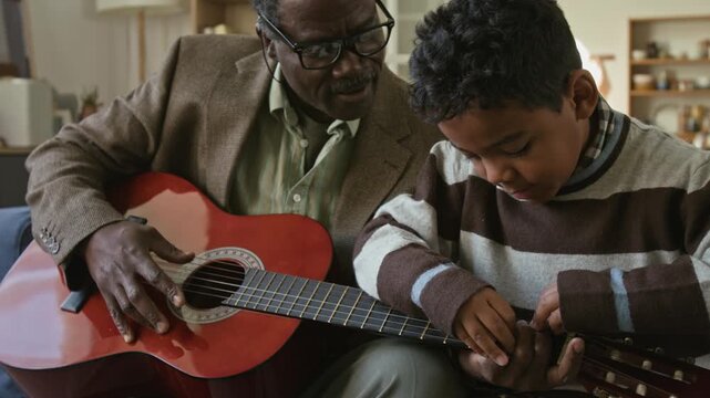 Focused little Black boy teaching granddad music chords holding down guitar strings together while bonding over creative hobby in living room on weekend