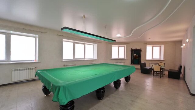 billiard room with a green-covered table. The room is lit by a rectangular overhead light.  Windows and seating are visible