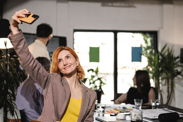 Business meeting with team members in a modern office during the afternoon, focusing on a woman taking a selfie while others engage in discussion