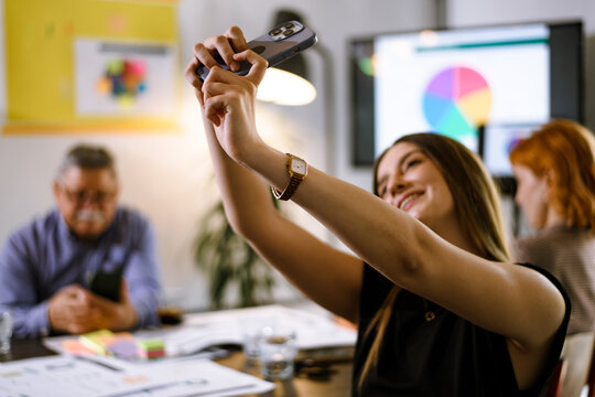 Group of colleagues engaging in a creative meeting while one person takes a selfie with a smartphone in a bright office space filled with colorful charts and documents