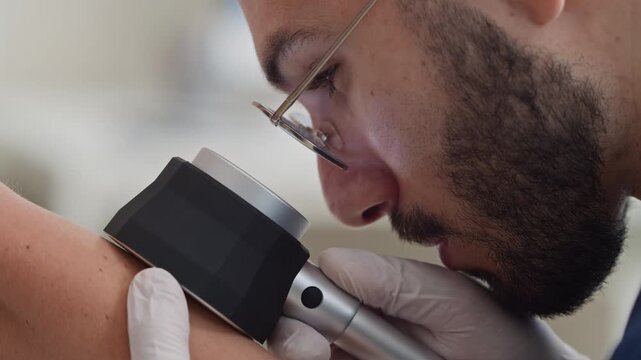 Close up shot of focused young Middle-Eastern male dermatologist in white sterile gloves using dermatoscope while examining carefully mole on unrecognizable patients arm in clinic
