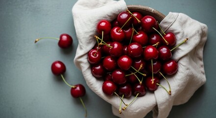 A bowl of fresh red cherries with stems on a white cloth and a gray background surface top down view