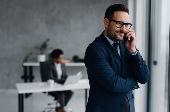 A businessman in a modern office standing near a window and using his smartphone and in the background, an African businesswoman is seated at a desk working on a laptop
 - Powered by Adobe