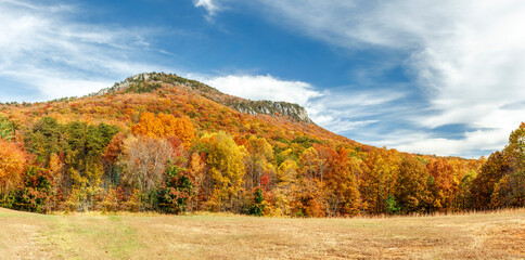 The steep, rocky cliffs of Cooks Wall, in Hanging Rock State Park in the North Carolina Sauratown Mountains, are surrounded by colorful fall foliage topped with a cloudy blue autumn sky.