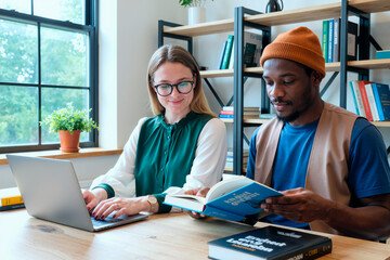 Young woman using laptop while Black young man reading book, both sitting at desk in modern office setting with bookshelves in background, collaborating on project