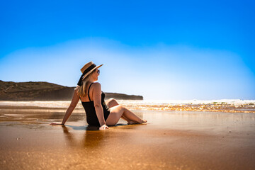Beautiful middle-aged woman sitting on seashore and sunbathing on sandy beach on Algarve coast in...