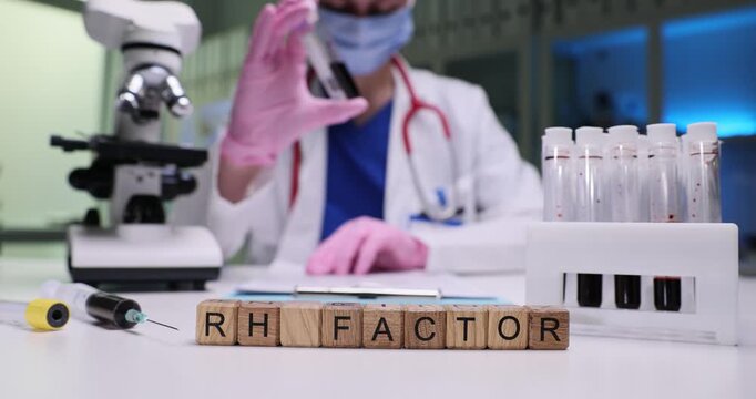 Wooden blocks spell words RH Factor on bright laboratory table with glassware. Masked doctor carefully lifts blood sample tube checking rhesus result