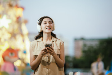 A smiling woman enjoying music with headphones while holding her smartphone, surrounded by bright decorative lights in a lively atmosphere.