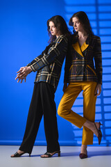 Stylish twin sisters showcase elegant fashion against a vibrant blue backdrop in a creative setting