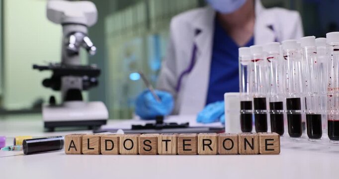 Wooden blocks spell word Aldosterone near blood test tubes. Laboratory doctor writes detailed hormone report studying patient sample by microscope
