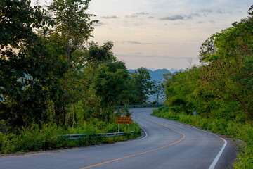 Countryside road with curve and slope on mountains and forest, Wonderful sunrise in the morning with golden yellow skyline and warm sunlight, Mueang Mai, Chae Hom District, Lampang province, Thailand.