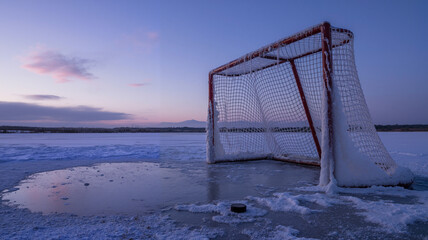Frozen ice hockey goal on a snowy lake at sunset with a hockey puck nearby