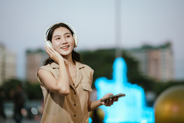 A smiling woman enjoying music with headphones while holding her smartphone, surrounded by bright decorative lights in a lively atmosphere.